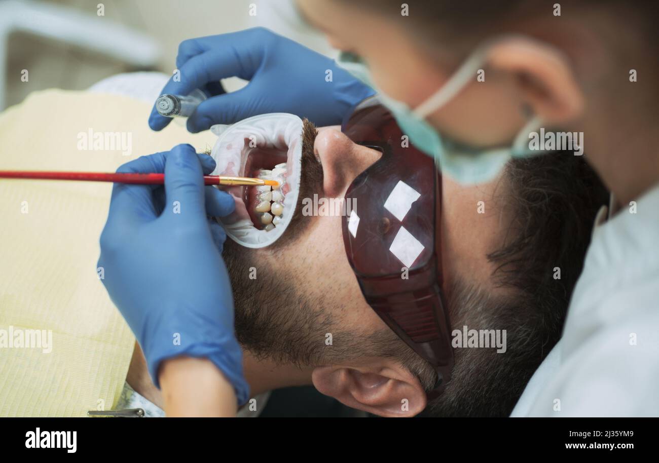 Un joli jeune homme souriant qui regarde l'appareil photo. Dentiste femme vérifiant les dents du patient avec miroir dans la clinique dentaire moderne. Examiner le jeune homme par le dentiste sur Banque D'Images