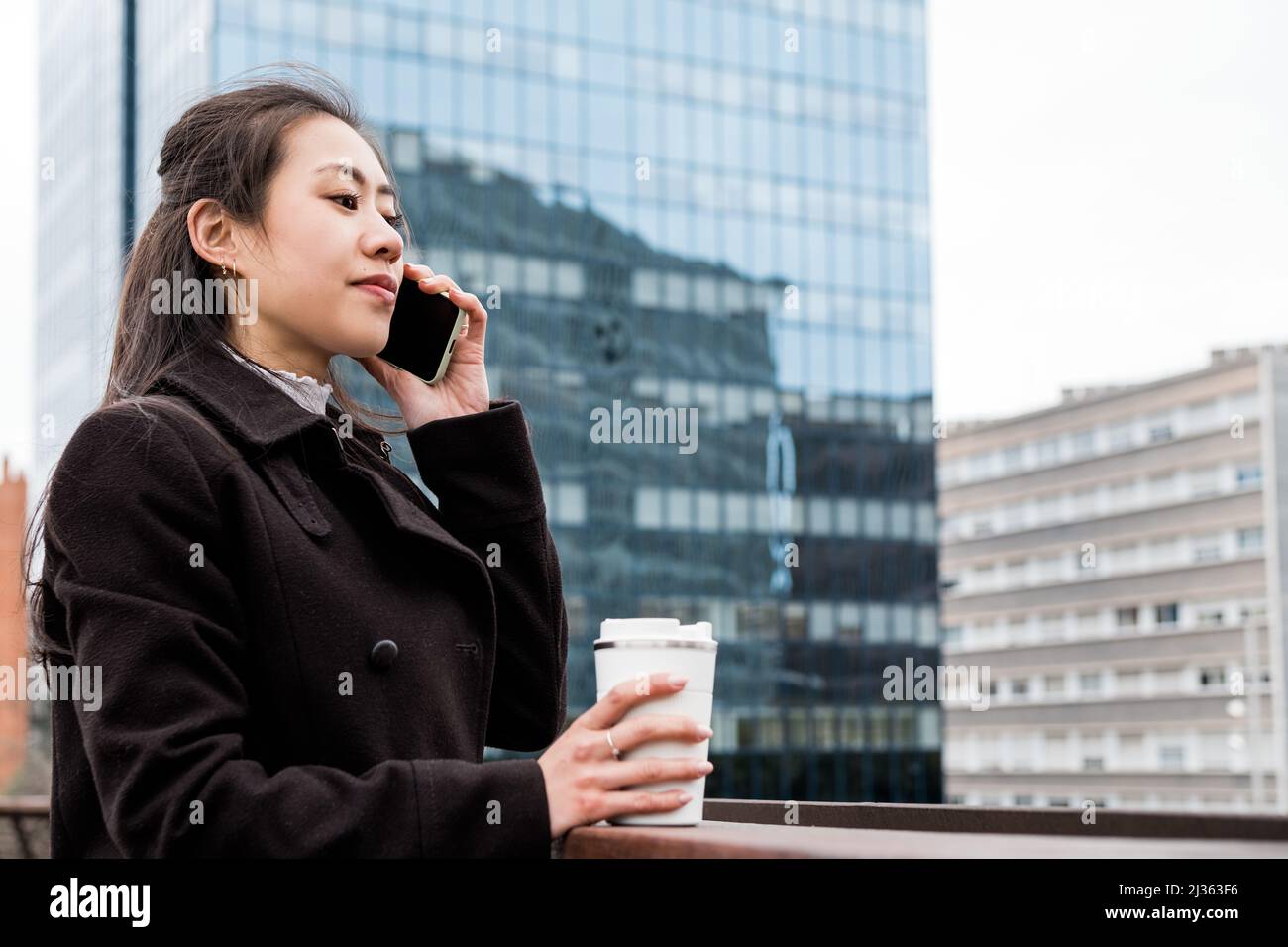 Femme chinoise gérant en manteau noir avec une tasse de café à emporter sans gaspillage. Elle est debout sur le pont et répond à un appel téléphonique le matin dans le centre-ville Banque D'Images