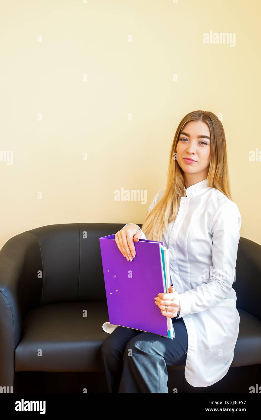 Portrait d'un jeune médecin caucasien avec des dossiers et des cheveux longs blonds portant un manteau blanc assis sur le canapé noir regardant l'appareil photo Banque D'Images