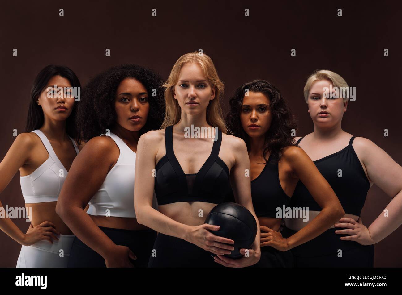 Cinq femmes en vêtements de sport debout ensemble. Groupe de jeunes femelles avec différents types de corps debout sur fond brun. Banque D'Images