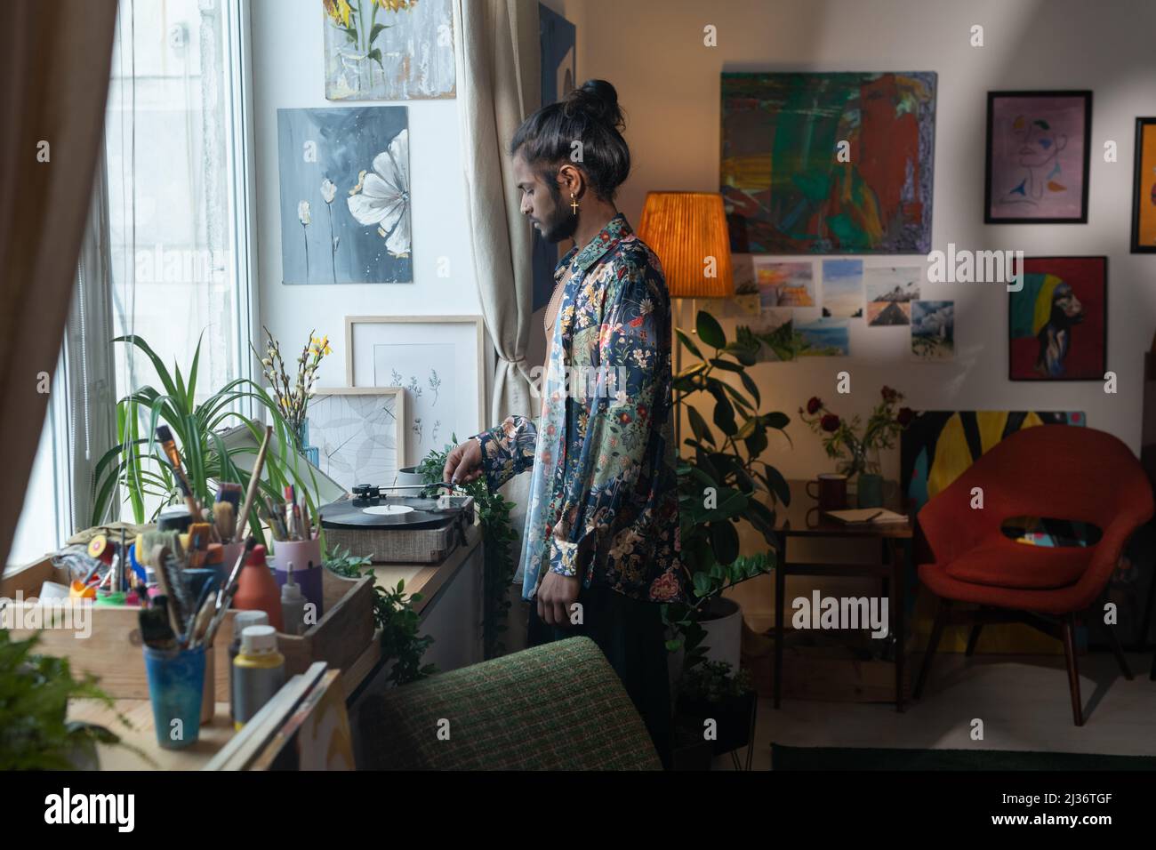 Jeune Indien créatif debout dans la salle de séjour décorée de diverses peintures tournant sur la musique sur vinyle lecteur de disques Banque D'Images