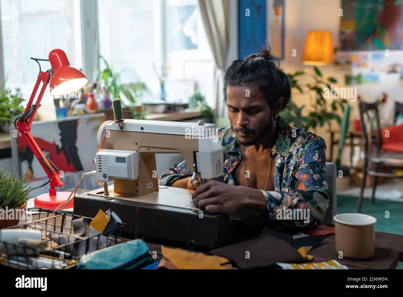 Portrait du jeune Indien créatif assis à une table dans le salon à la maison en utilisant la machine à coudre pour faire des vêtements neufs Banque D'Images