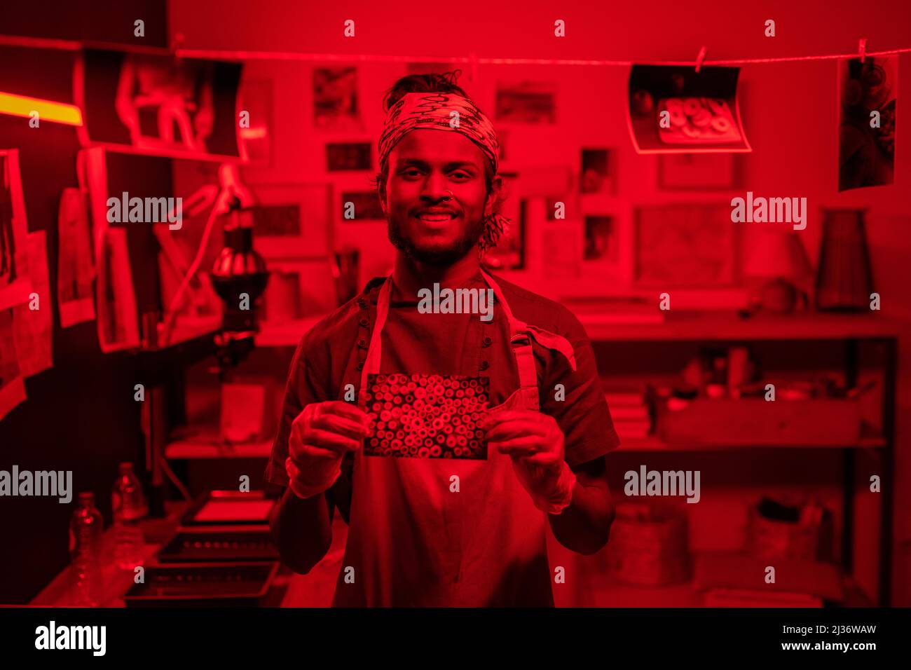 Joyeux jeune Indien portant un tablier et des gants de protection debout dans une chambre noire démontrant la photo développée et souriant à la caméra Banque D'Images