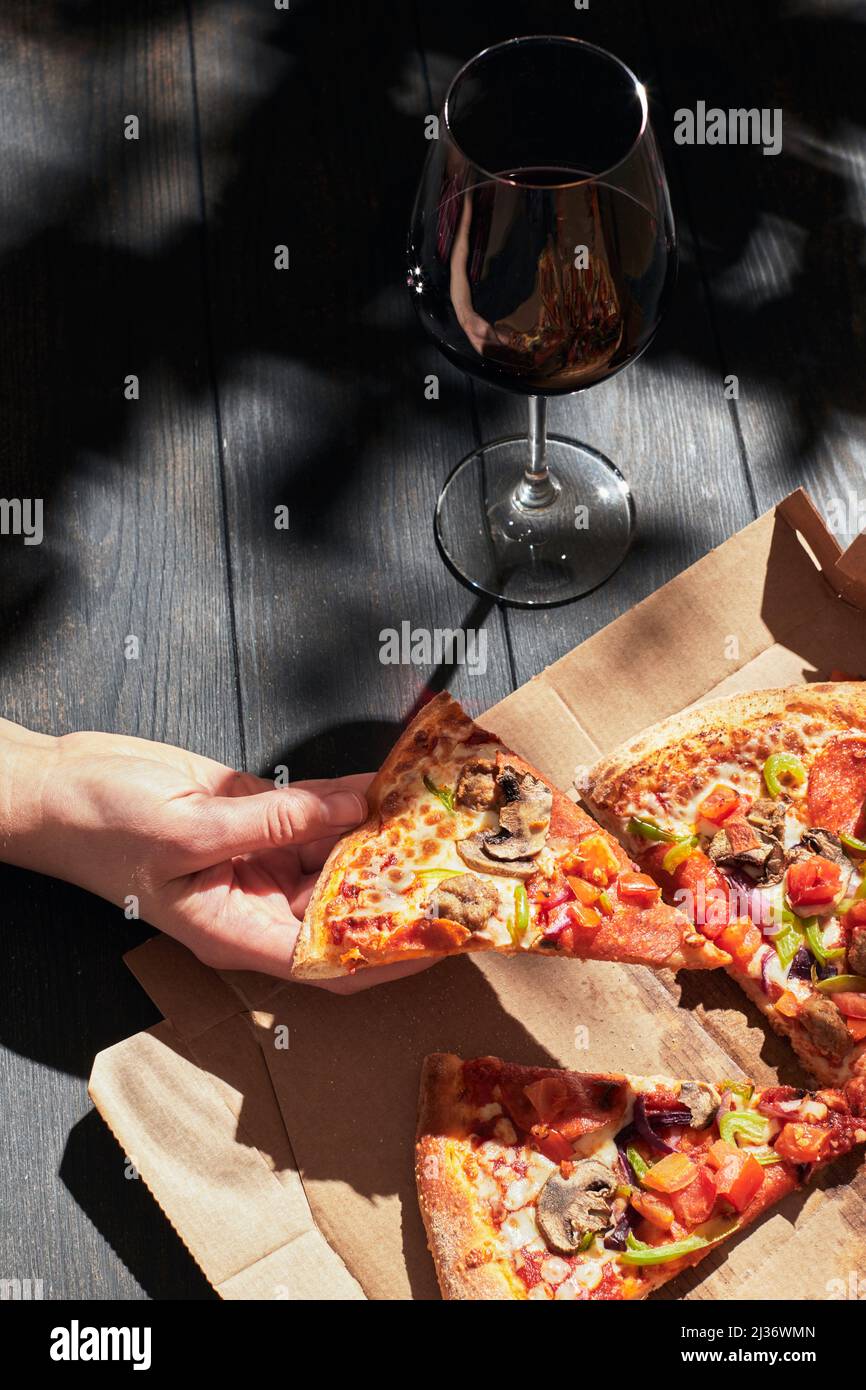 La main d'une femme prend une tranche de pizza par temps ensoleillé. Pizza dans une boîte et un verre de vin. Le concept de livraison de nourriture. Photo verticale. Banque D'Images