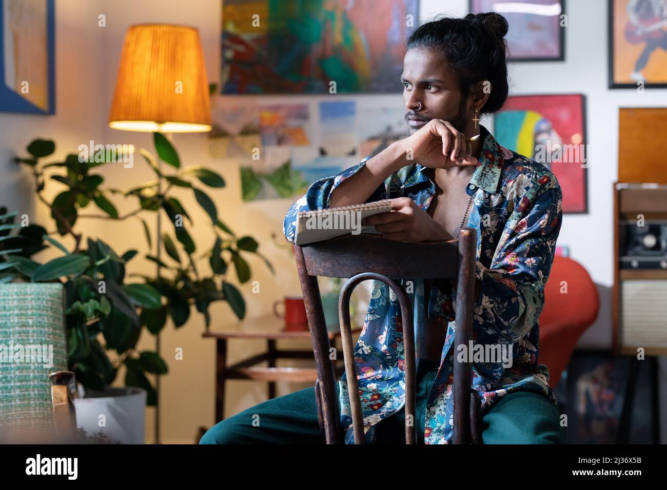 Portrait moyen d'un jeune Indien élégant portant une tenue décontractée assis sur une chaise en bois tenant un carnet à croquis et un crayon décidant de quoi dessiner Banque D'Images