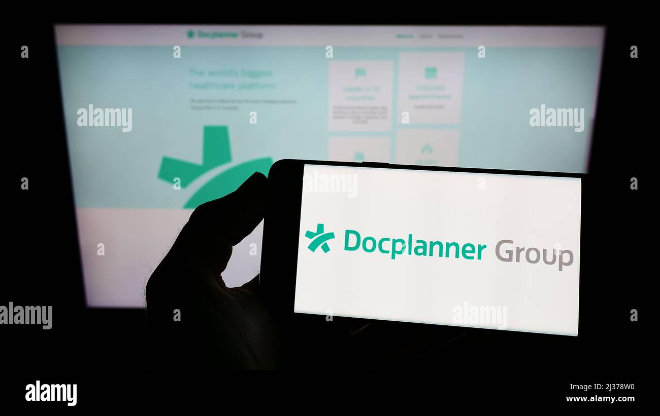 Personne tenant un téléphone portable avec le logo de la société polonaise de soins de santé DocPlanner Group à l'écran en face de la page Web d'affaires. Mise au point sur l'affichage du téléphone. Banque D'Images