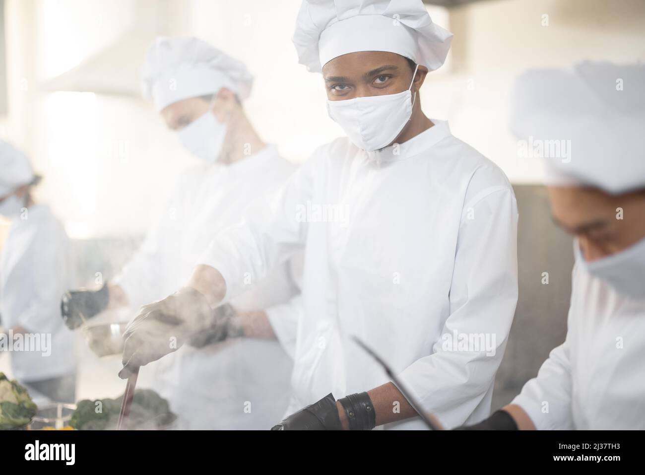 Une équipe multiraciale de cuisiniers cuisiniers en uniforme blanc dans la cuisine. Chef asiatique avec cuisine latine friture de viande, les gars européens cuisinent sur fond. Cuisiniers portant des masques et des gants de protection Banque D'Images