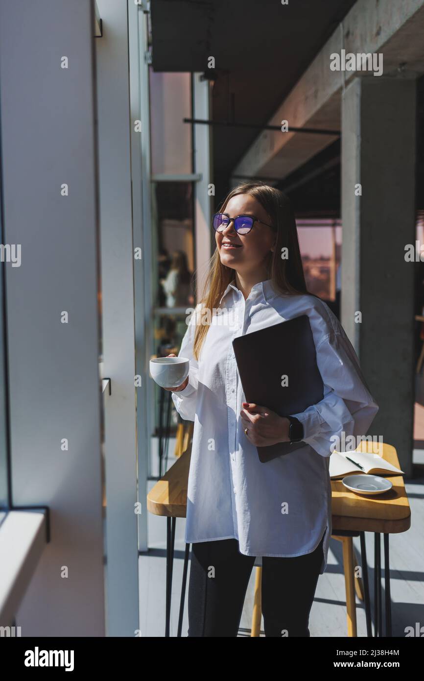 Femme d'affaires dans une chemise blanche et des lunettes avec un  ordinateur portable dans les mains au bureau près de la fenêtre. Femme  d'affaires moderne dans des vêtements élégants. Freelance Photo Stock -