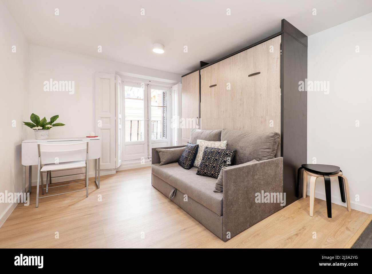 canapé-lit en tissu gris avec coussins bleus et blancs, balcon avec volets en bois, table à manger pliante et chaise assortie, armoire intégrée dans le vacatio Banque D'Images