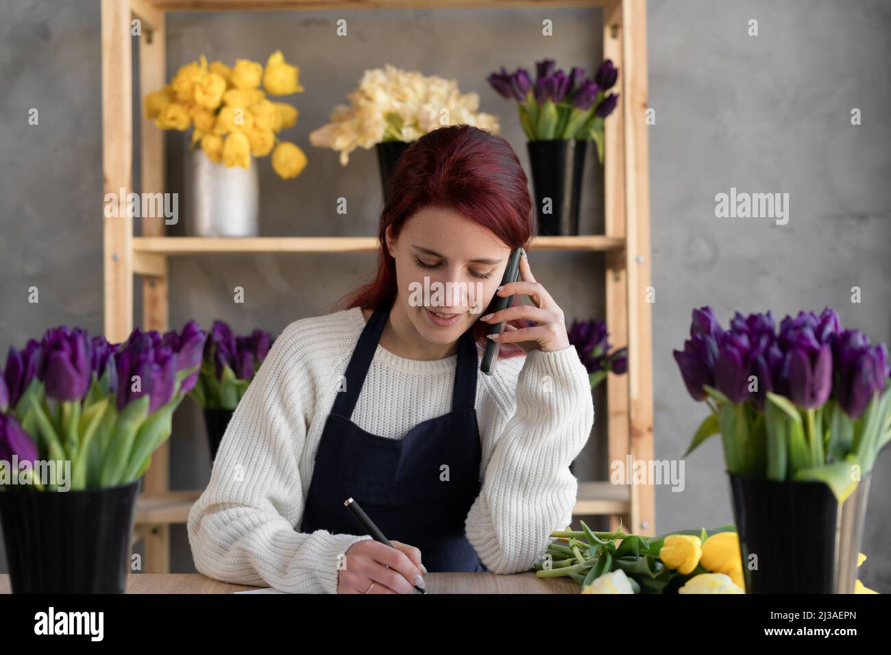 femme caucasienne dans un tablier. La femme propriétaire d'entreprise de fleur prend une commande en ligne pour un bouquet de fleurs par téléphone et écrit dans un carnet le Banque D'Images