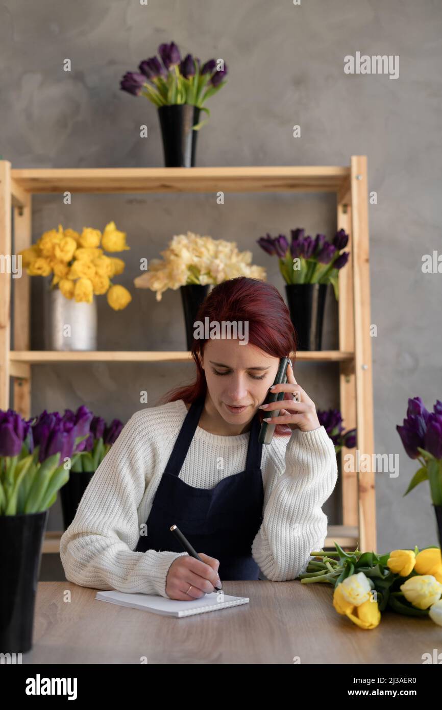 Portrait d'une dame du caucase dans un tablier. La femme propriétaire d'entreprise de fleur prend une commande en ligne pour un bouquet de fleurs par téléphone et écrit vers le bas dans un Banque D'Images