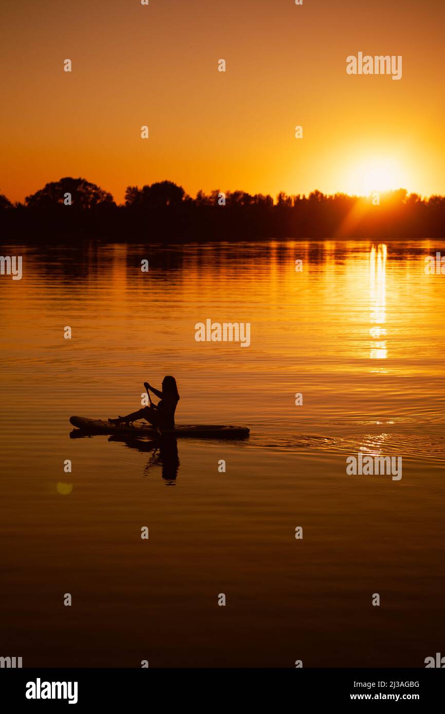 Silhouette de dame assise sur le plan supérieur en pagayant dans les mains sur le lac avec le soleil se coucher derrière l'horizon en arrière-plan couvrant la surface de l'eau avec de l'orange Banque D'Images