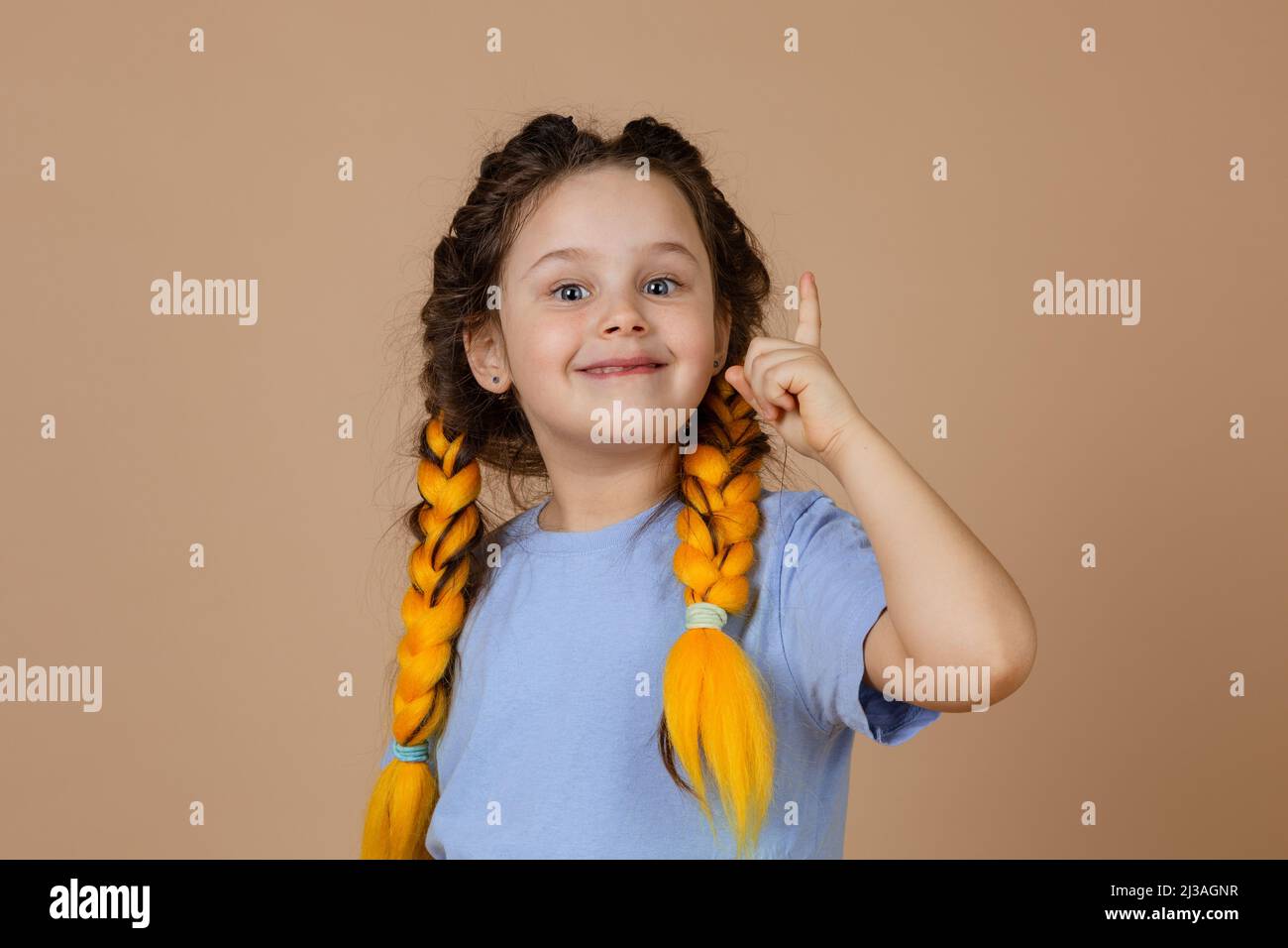 Jeune fille de génie avec des tresses jaunes de kanekalon montrant le doigt de point indiquant une grande idée apparaissant dans la tête sur fond beige. Eureka Banque D'Images