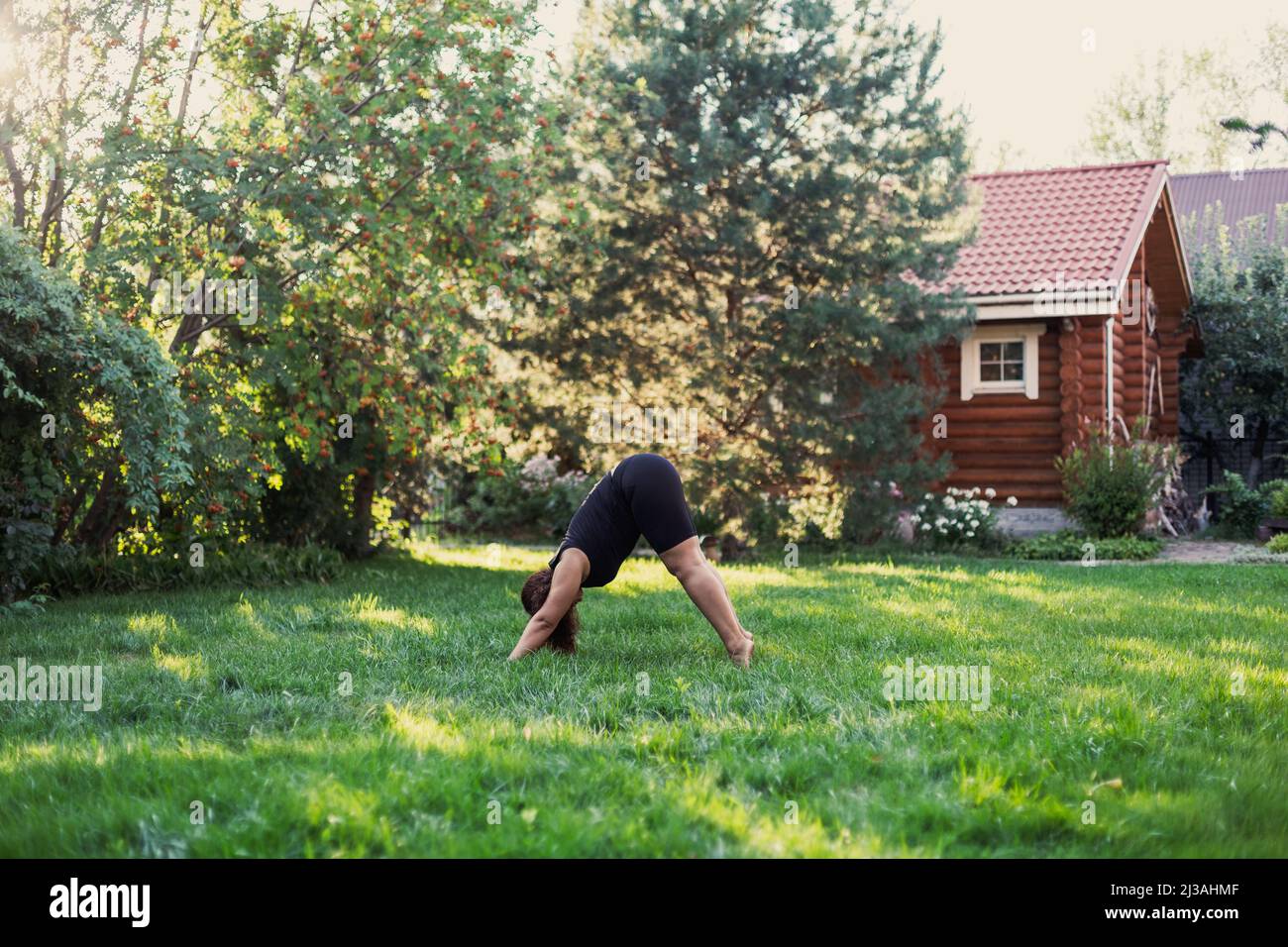 Femelle sur-pondérée faisant du yoga debout en position triangle sur l'arrière-cour du chalet avec maison en bois et arbres en arrière-plan. Corps positif Banque D'Images
