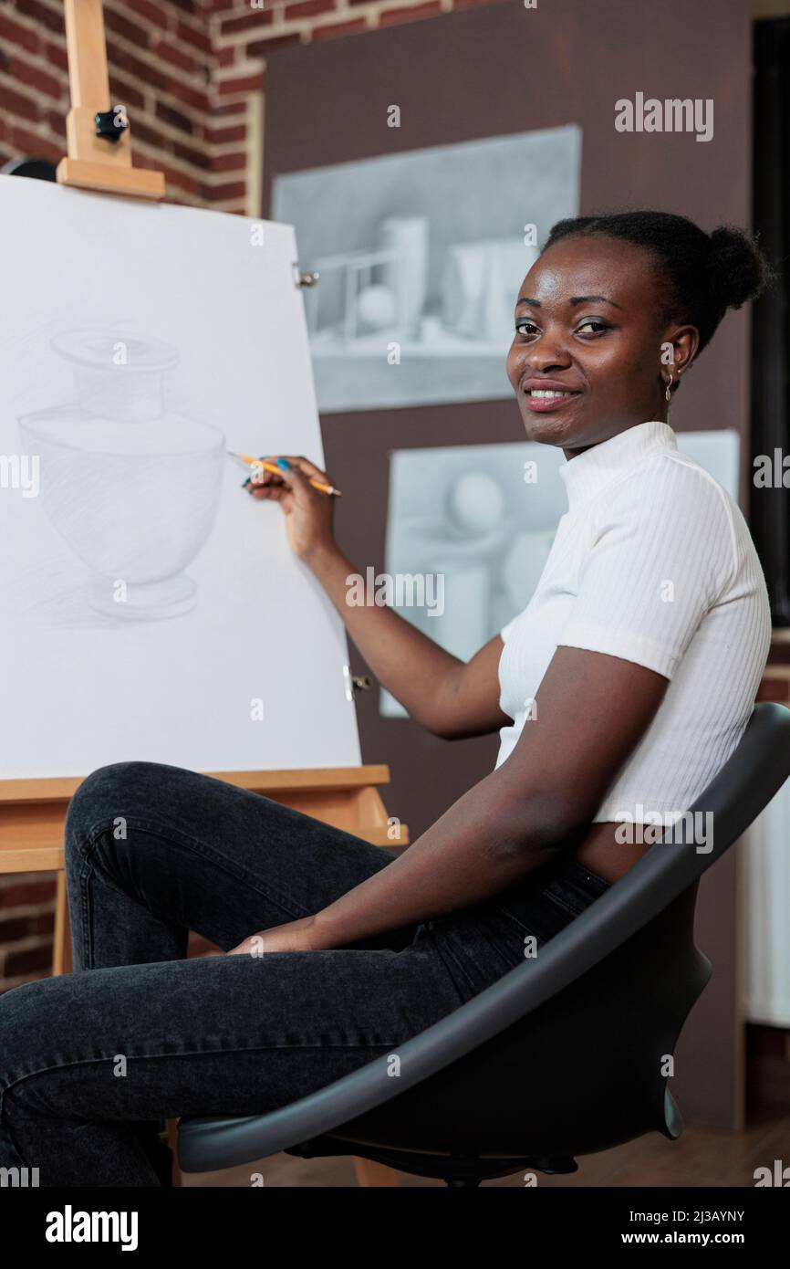 Portrait d'un étudiant souriant qui s'attlait à un cours de dessin en profitant d'une leçon de croquis en studio de créativité. Peintre femme esquissant un modèle de vase sur toile blanche travaillant à la technique graphique. Concept créatif Banque D'Images