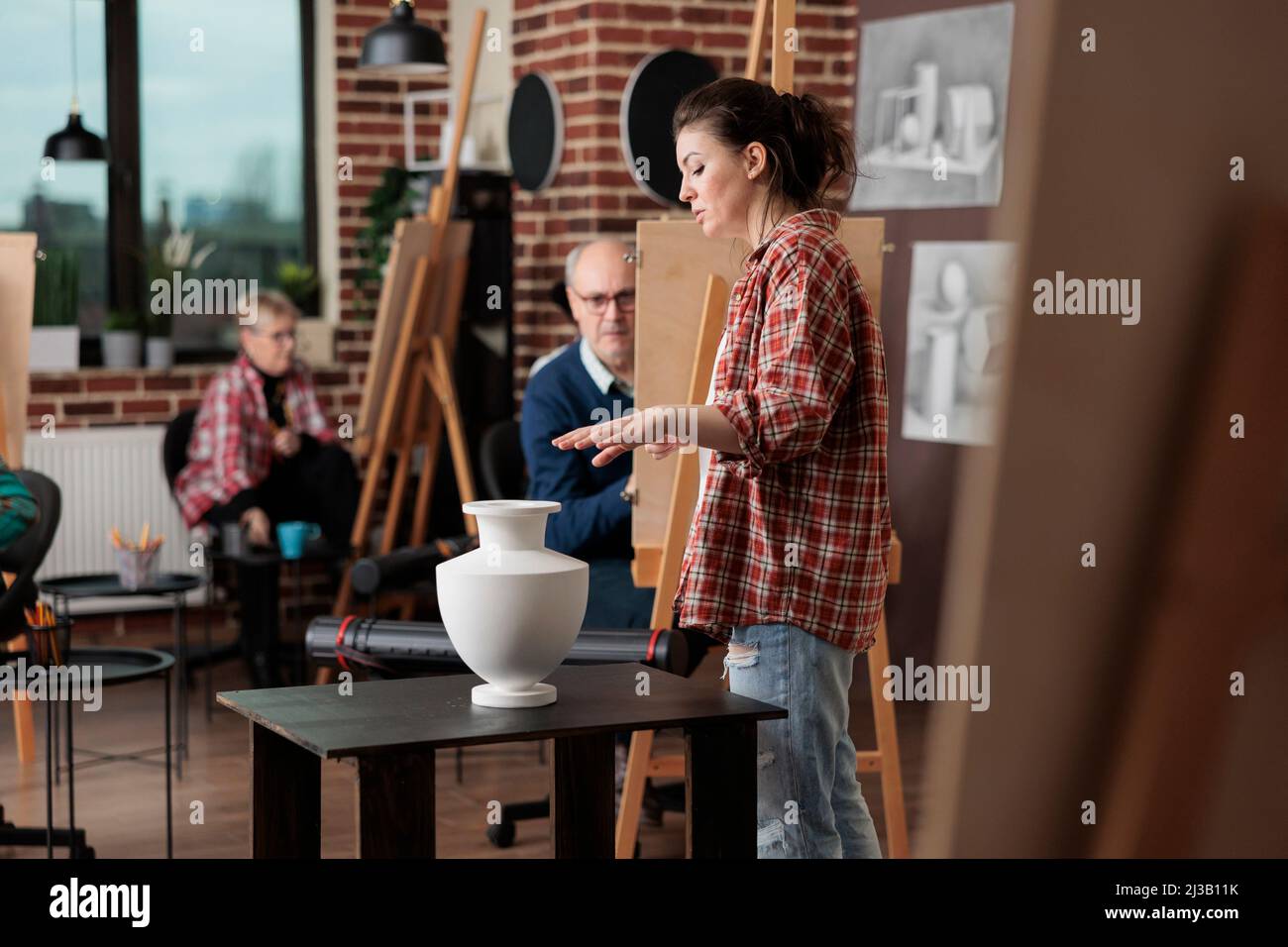 Jeune professeur de peintre supervisant des cours d'art expliquant la technique graphique à divers étudiants en studio de créativité. Personnes multiethniques esquisse vase modèle sur toile d'apprentissage artistique compétence Banque D'Images