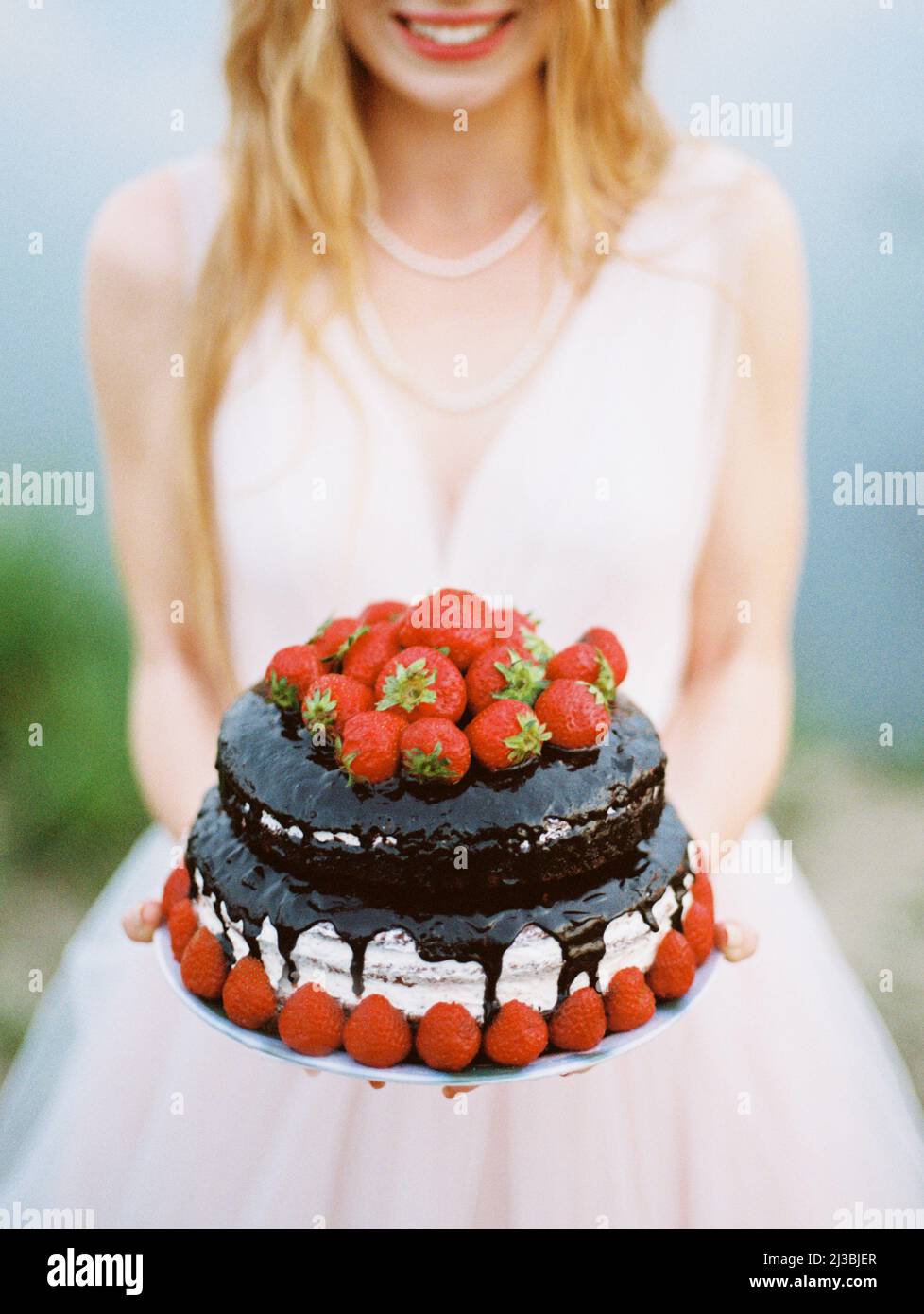 Gâteau de chocolat de mariage gros plan avec des fraises dans les mains de la mariée Smilimg en robe de mariage rosé. Mariage rustique en plein air. Banque D'Images