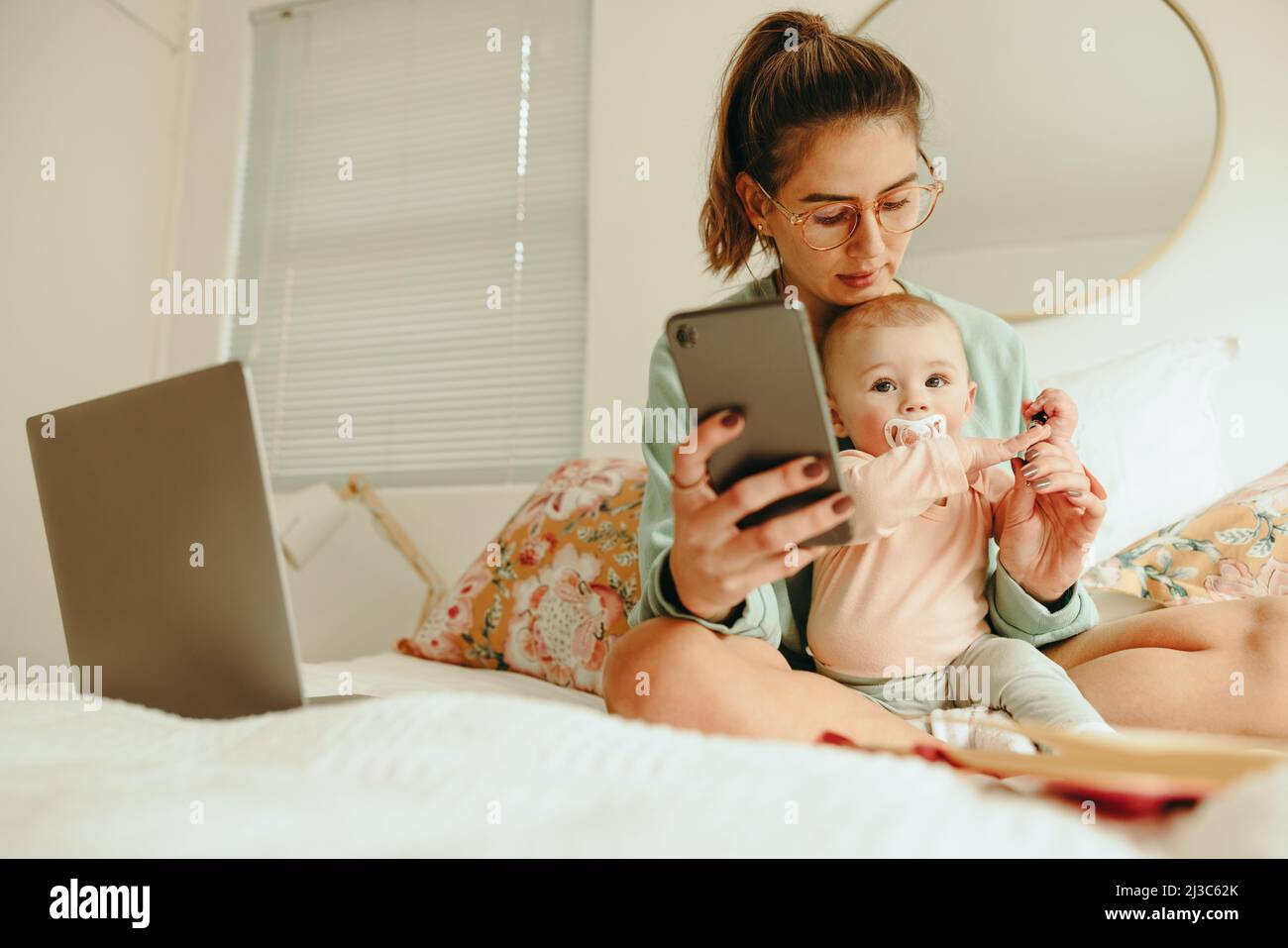 Équilibre entre la maternité et le travail. Une nouvelle maman prend un stylo de son bébé tout en travaillant sur un lit. Multitâche mère seule essayant de lire un message texte whi Banque D'Images
