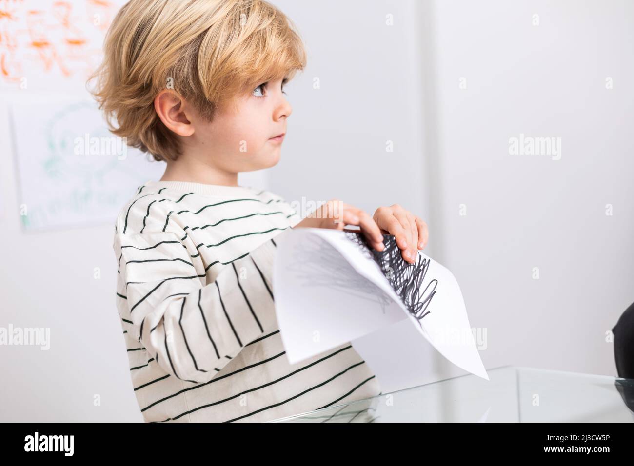 Mignon garçon aux cheveux blonds papier blanc pliant avec des coups noirs tout en étant assis dans la pièce claire pendant le rendez-vous avec le psychologue Banque D'Images