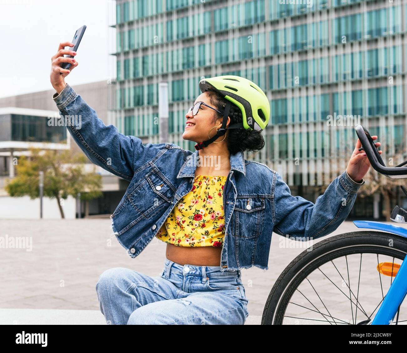 Femme colombienne souriante en denim et casque de protection emportant un selfie sur un smartphone tout en prenant une pause à vélo dans la rue de la ville Banque D'Images