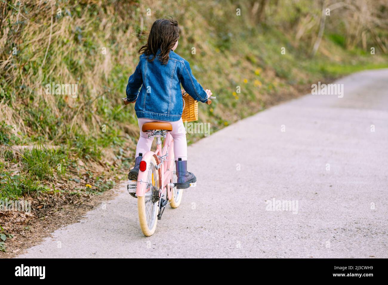 Vue de bach du corps entier d'un enfant méconnaissable dans une veste en denim à vélo sur la piste le long du parc vert le jour de l'été Banque D'Images