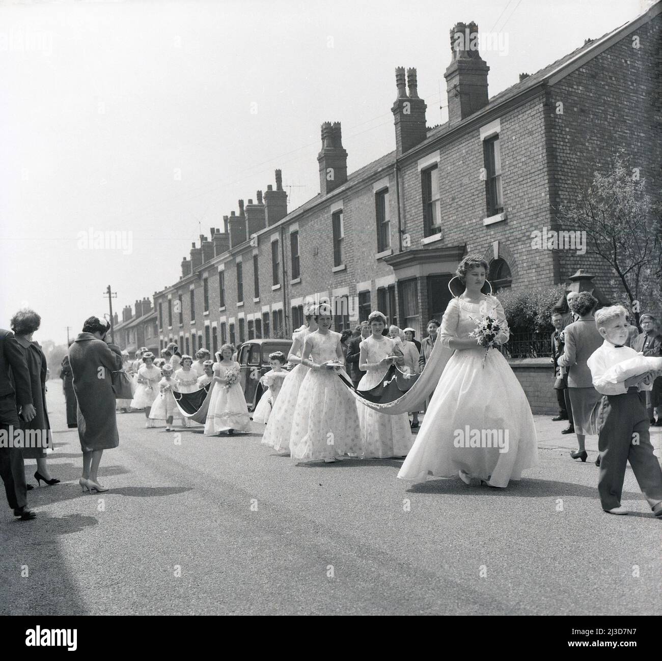 1961, historique, procession du jour de mai, deux jeunes filles en longues robes tiennent le train lourd de la nouvelle Reine de mai couronnée de la ville, alors qu'ils marchent le long d'une rue de maisons victoriennes en terrasse à Stockport, Manchester, Angleterre, Royaume-Uni. Banque D'Images