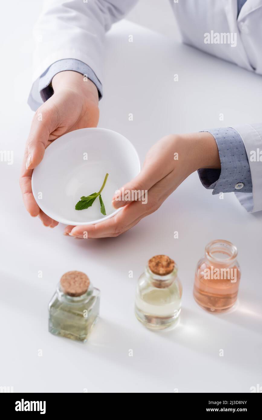 vue rognée de l'assistant de laboratoire tenant le bol avec des feuilles de menthe près des bouteilles dans le laboratoire Banque D'Images