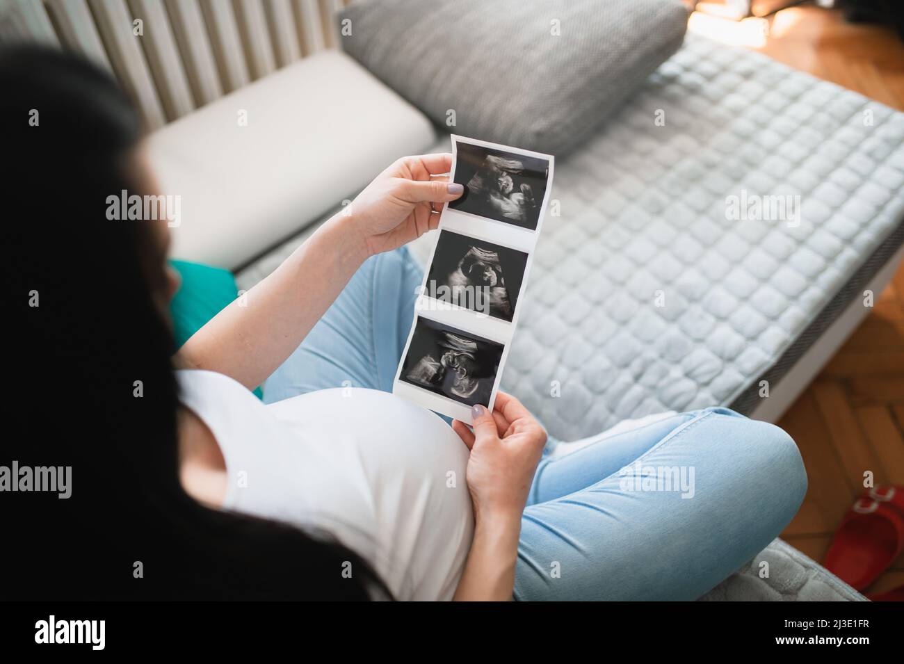 Une femme enceinte caucasienne à cheveux noirs regarde des images de bébé à ultrasons. Elle tient du papier à ultrasons devant son ventre de grossesse. Une femme est assise. Banque D'Images