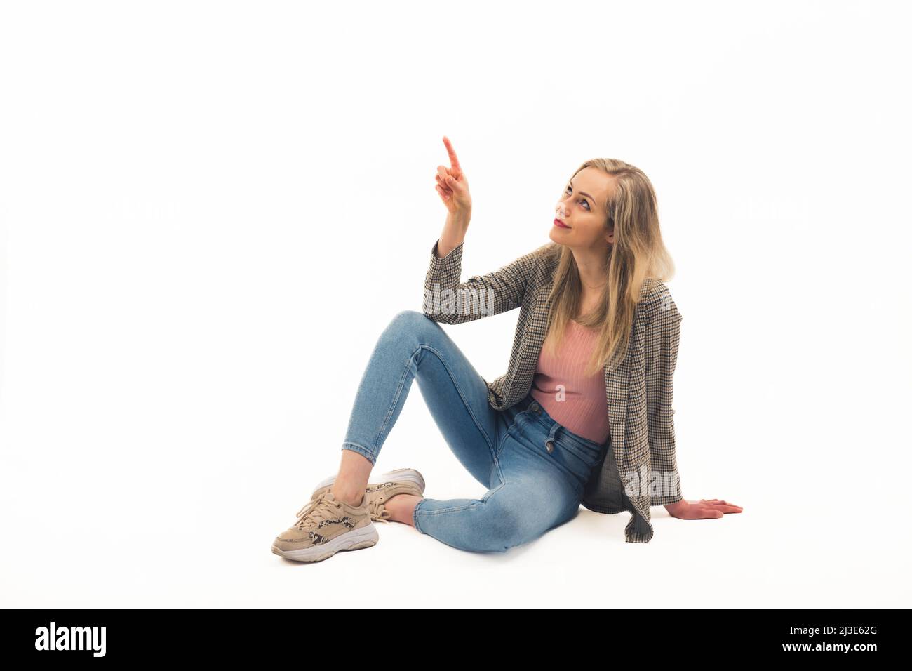 Agréable blanche blonde fille assise sur le sol et avec un doigt pointant vers le haut plein plan studio tourné blanc arrière-plan copie espace. Photo de haute qualité Banque D'Images