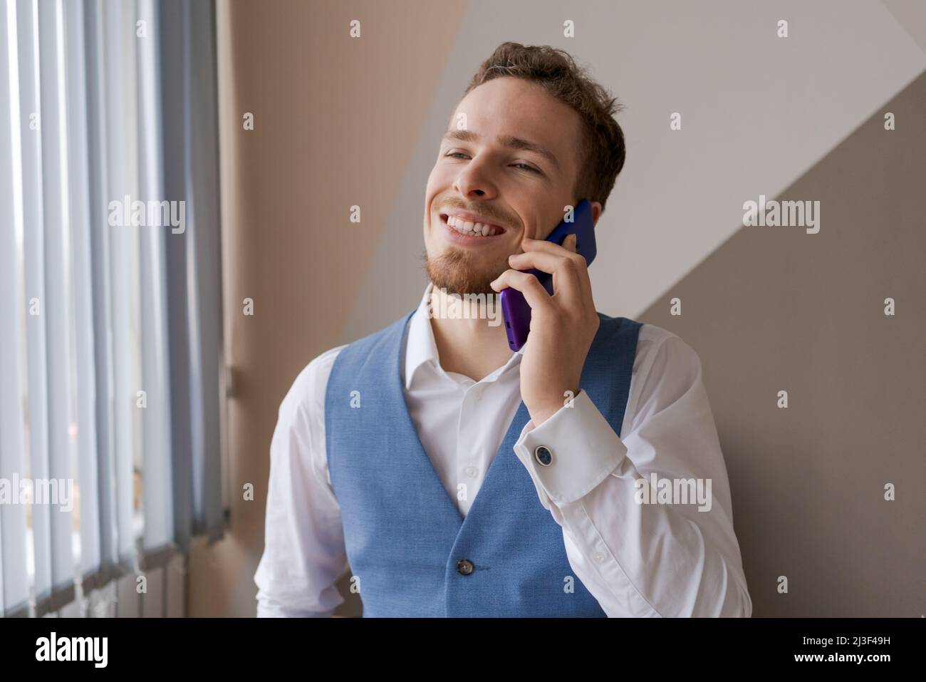 Agréable beau barbu homme en chemise et gilet parler au téléphone, commander un taxi ou la livraison de nourriture au bureau au travail. Un jeune homme d'affaires caucasien souriant tient un appel avec des clients partenaires. Banque D'Images