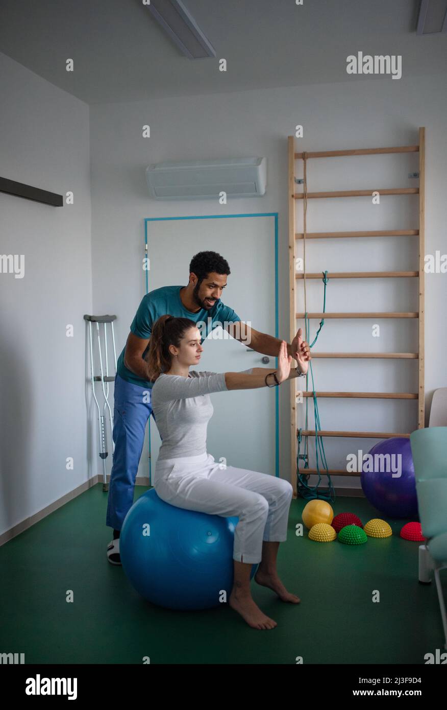 Un jeune physiothérapeute masculin fait de l'exercice avec une jeune femme patiente sur le ballon dans une salle de physique Banque D'Images
