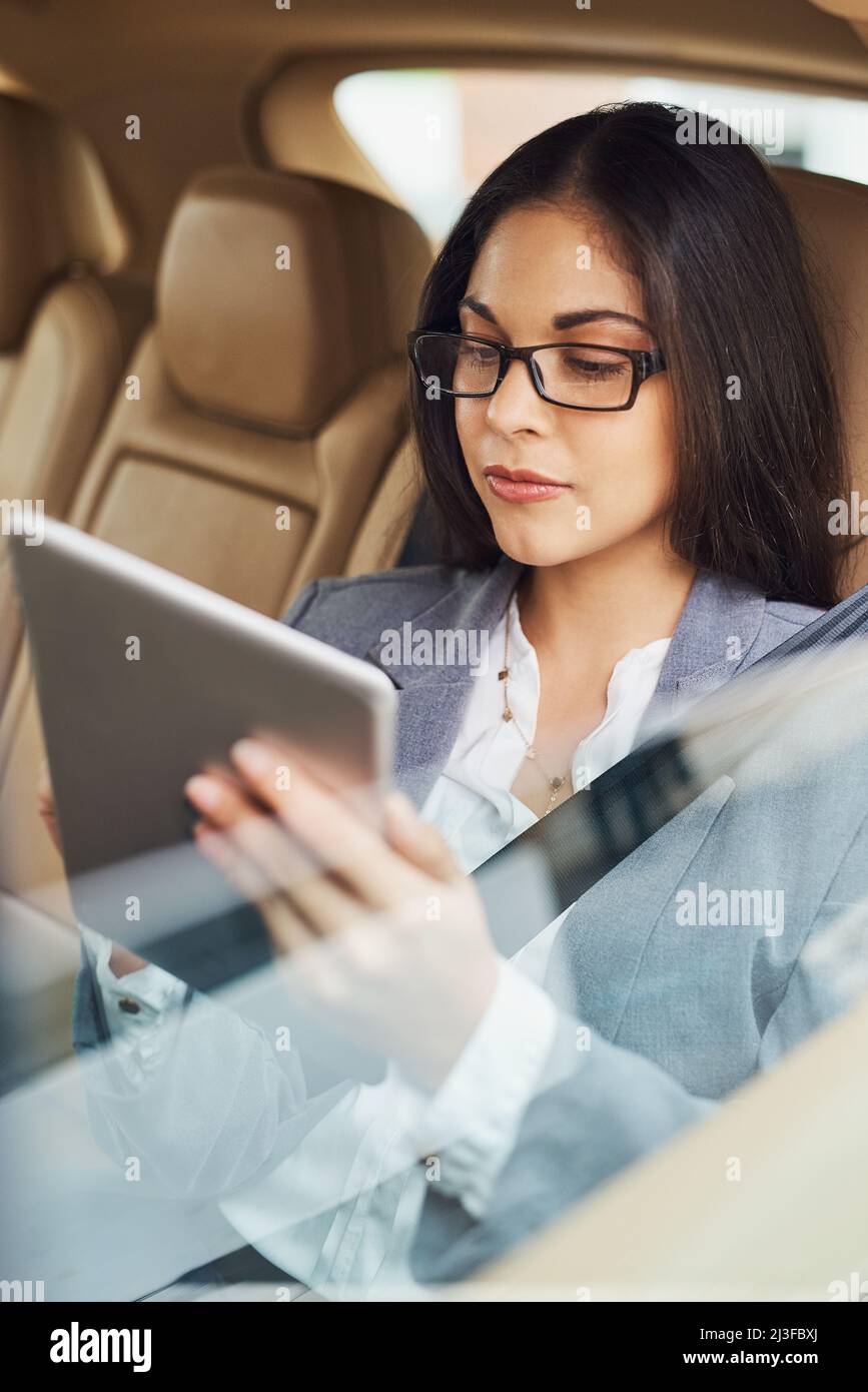 Obtenir une mise à jour de votre entreprise en déplacement. Prise de vue d'une jeune femme d'affaires utilisant sa tablette numérique tout en étant assise dans le siège arrière d'une voiture. Banque D'Images