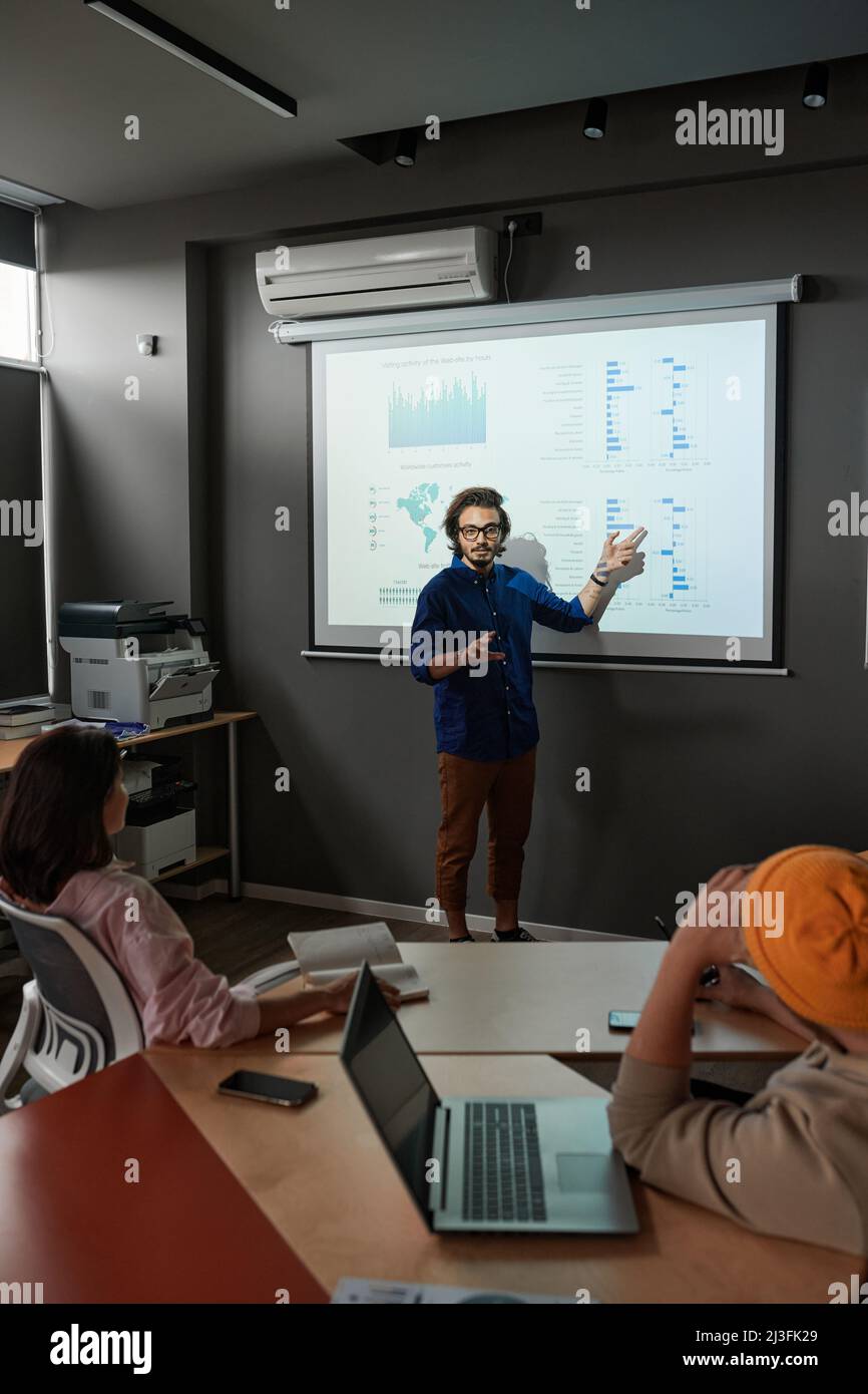 Jeune analyste de race mixte pointant sur l'écran de projection tout en présentant des statistiques lors d'une réunion du personnel Banque D'Images