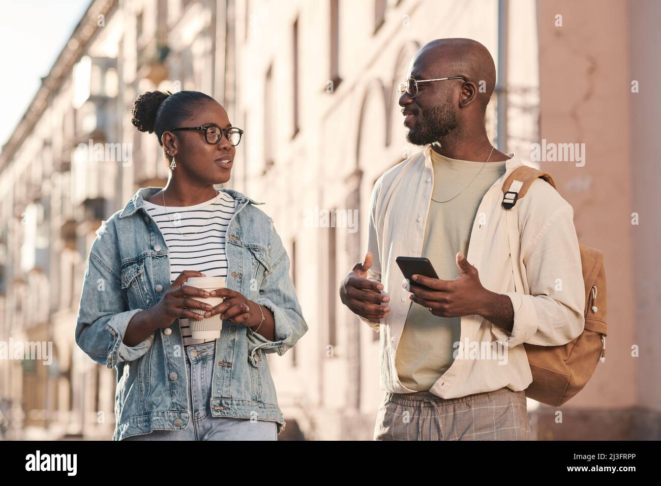 Jeunes amis afro-américains positifs en costume décontracté buvant du café et parlant tout en marchant ensemble en ville Banque D'Images