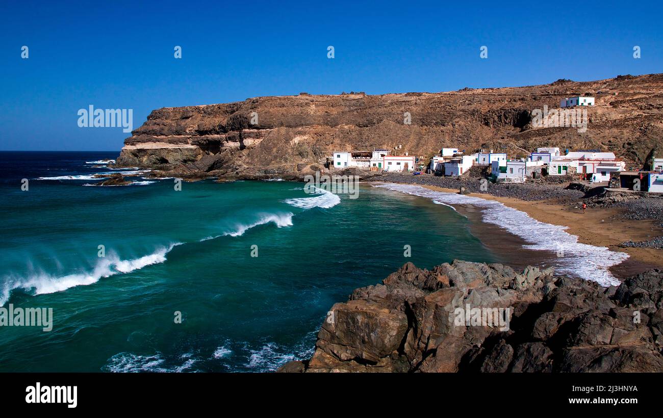 Espagne, îles Canaries, Fuerteventura, côte ouest, El Puertito de los Molinos, petit village côtier, vue sur les maisons, rochers en premier plan à droite, vert de mer avec surf, dans les rochers de fond de la baie, ciel bleu sans nuages Banque D'Images