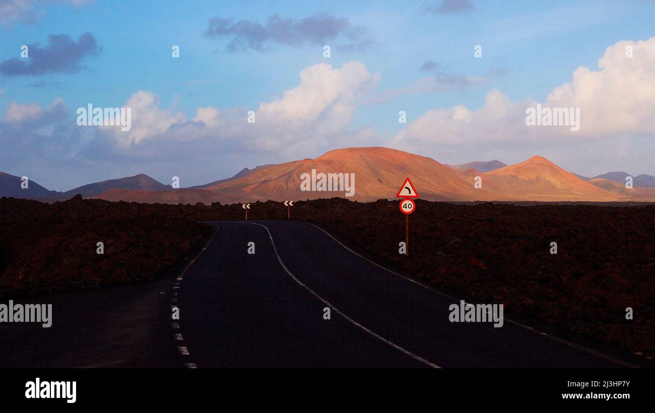 Îles Canaries, Lanzarote, île volcanique, parc national de Timanfaya, paysages volcaniques, Une route asphaltée à l'ombre mène au parc national, cratère volcanique rouge-brun au milieu, ciel bleu, nuages gris et blanc, panneau de signalisation « 40 », panneau de signalisation « Sharp Left Curve » Banque D'Images