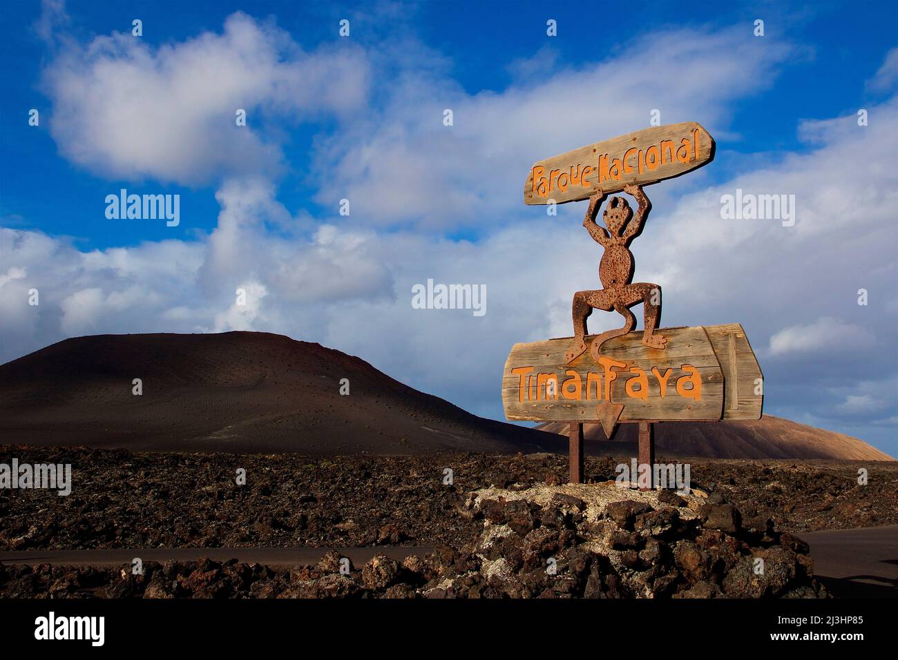 Îles Canaries, Lanzarote, île volcanique, parc national de Timanfaya, paysages volcaniques, route d'entrée au parc national, panneau d'entrée en bois, figure du monument de Lanzarote, le diable de feu est sur le panneau 'Timanfaya', avec ses mains levées il tient le panneau avec l'inscription 'Parque Nacional ', ciel bleu avec des nuages blancs, la roche de lave se trouve autour du panneau Banque D'Images