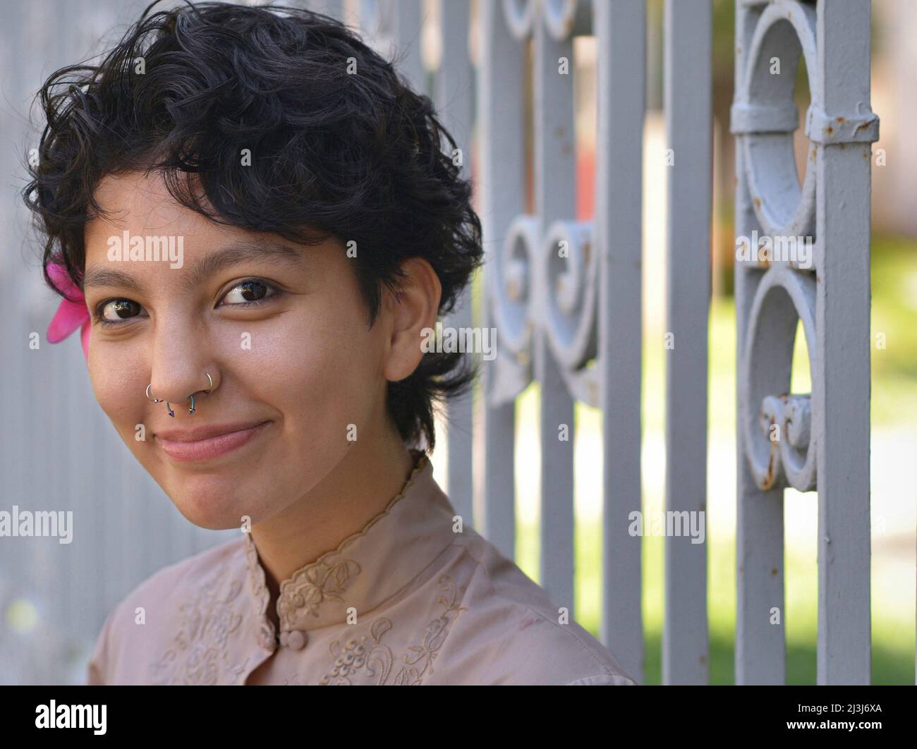 Jeune femme très sensuelle Latina avec de grands yeux bruns et une fleur dans son petit cheveux noir et courbé regarde le spectateur devant une clôture en fer forgé. Banque D'Images