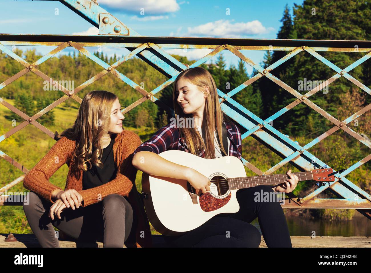 Deux sœurs s'assoient sur un pont de parc, dont une avec une guitare acoustique, en écoutant de la musique ensemble; Edmonton, Alberta, Canada Banque D'Images