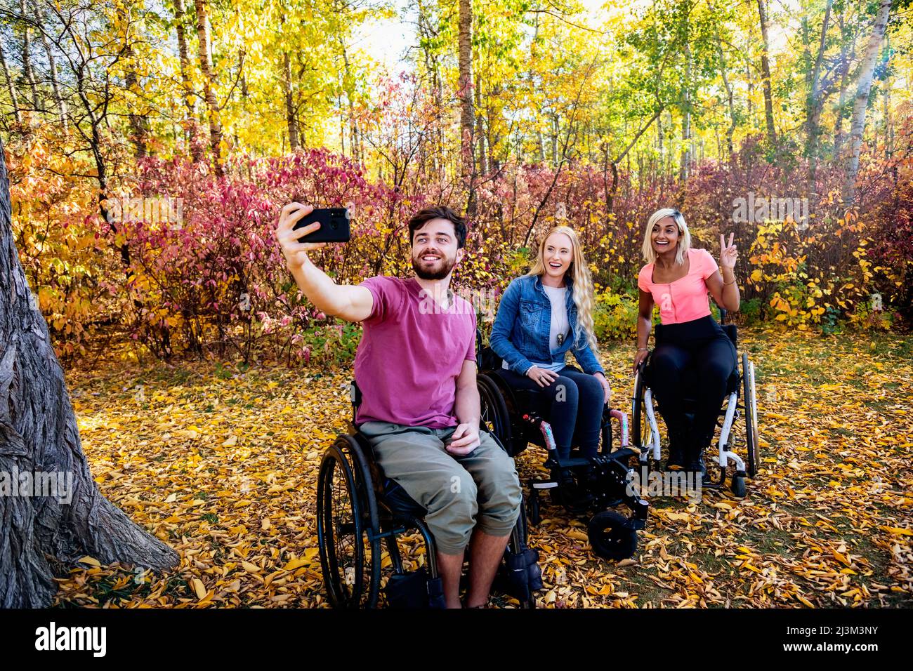 Jeunes hommes et femmes paraplégiques en fauteuil roulant prenant un autoportrait avec un smartphone dans un parc lors d'une belle journée d'automne Banque D'Images