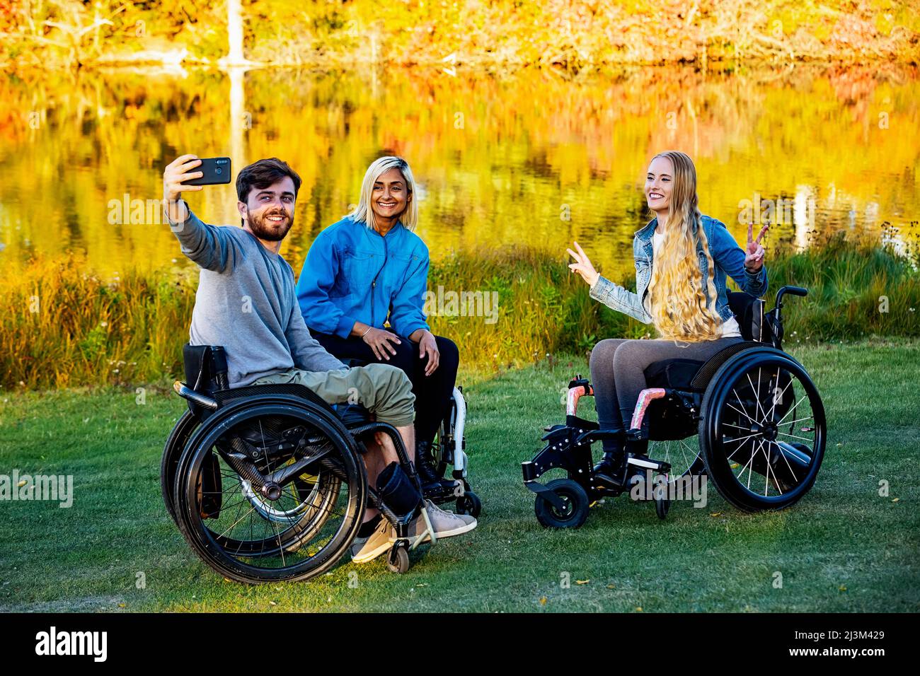 Jeunes hommes et femmes paraplégiques en fauteuil roulant prenant un autoportrait avec un smartphone dans un parc lors d'une belle journée d'automne Banque D'Images