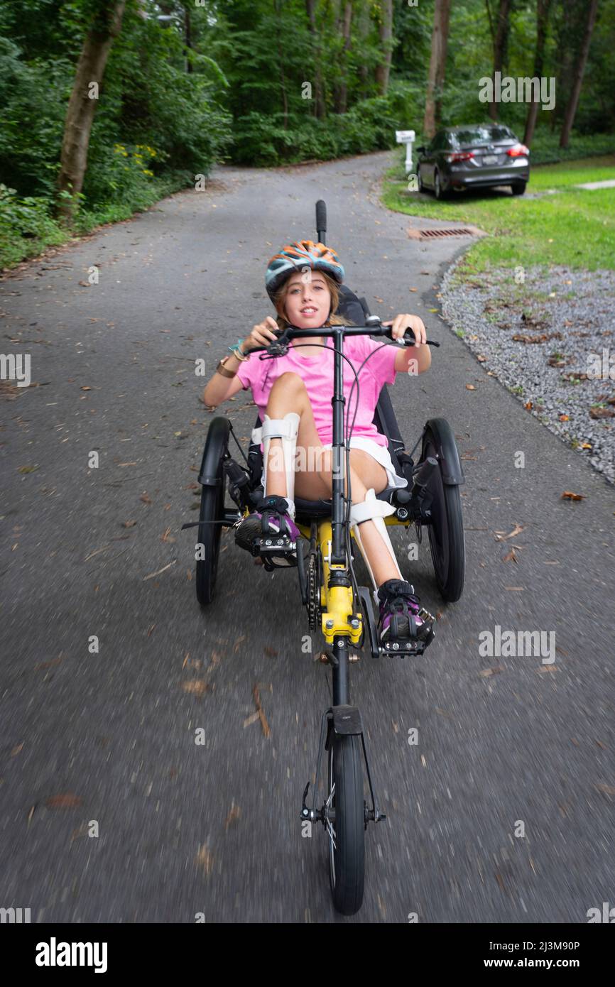 Fille avec la dystrophie musculaire congénitale d'Ullrich à cheval sur son vélo de trike allongé dans la rue près de sa maison Banque D'Images