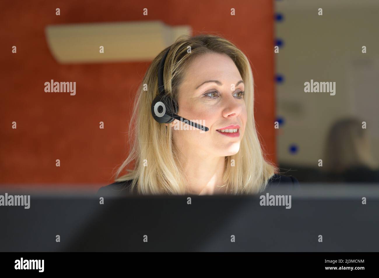 La réceptionniste ou l'opérateur du centre d'appels prend un appel téléphonique sur un casque vu par le haut de son ordinateur alors qu'elle regarde de côté avec un sourire Banque D'Images