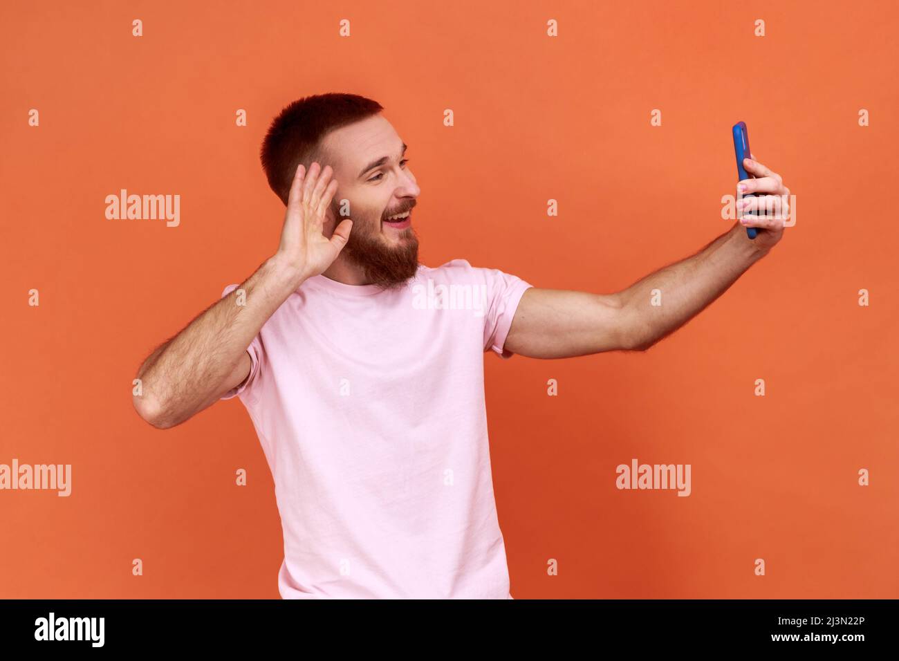 Portrait de l'homme barbu qui parle lors d'un appel vidéo et qui agite le geste de bonjour, qui a une conversation en ligne sur un téléphone mobile, en portant un T-shirt rose. Studio d'intérieur isolé sur fond orange. Banque D'Images