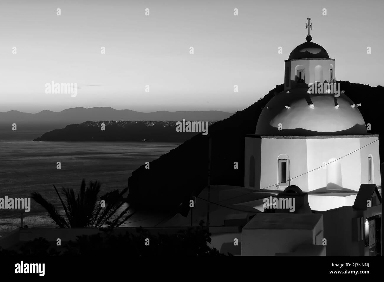 Vue sur une magnifique église orthodoxe blanchie à la chaux et sur Oia Santorini en arrière-plan, tandis que le soleil se couche de façon spectaculaire en noir et blanc Banque D'Images