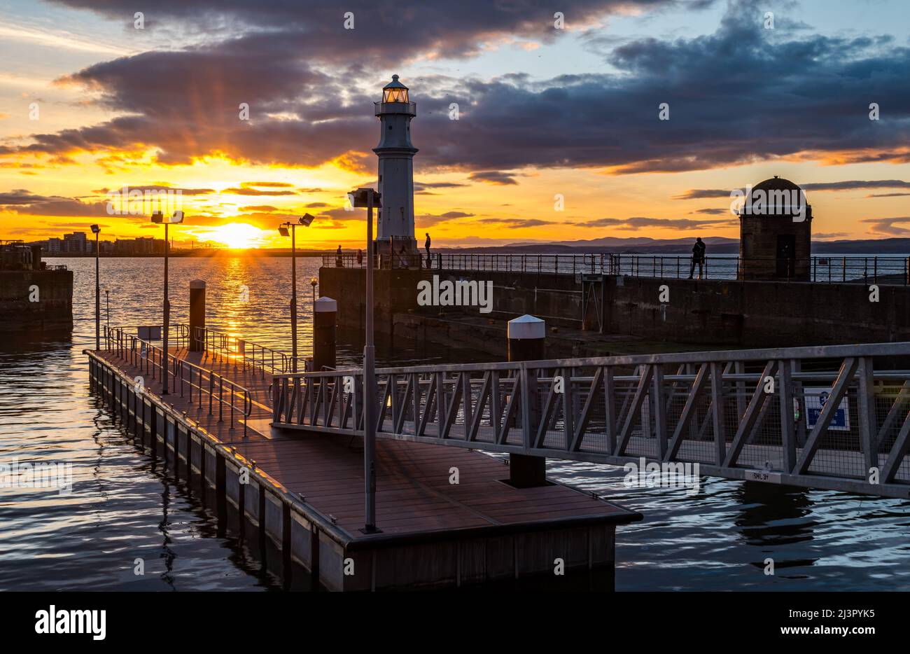 Édimbourg, Écosse, Royaume-Uni, 9th avril 2022. Météo au Royaume-Uni : coucher du soleil au port de Newhaven : le soleil se couche sur le phare de Firth of Forth et de Newhaven lors d'une journée de printemps froide mais ensoleillée tandis que les gens marchent sur le mur du port. Banque D'Images