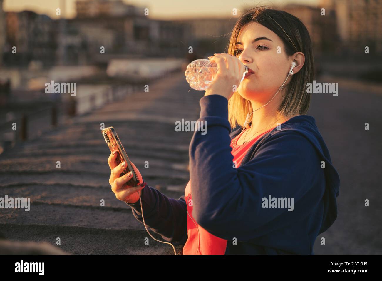 Une femme aux courbes, portant des écouteurs, boit de l'eau et fait une pause après son entraînement au coucher du soleil Banque D'Images