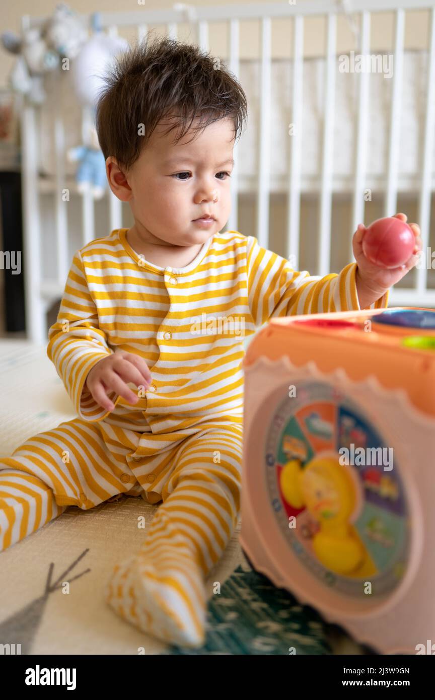 bébé garçon de 8 mois de course mixte jouant avec une boîte d'activité dans la chambre à coucher portant un pyjama jaune tout en étant assis sur le sol recouvert d'un jeu en caoutchouc Banque D'Images