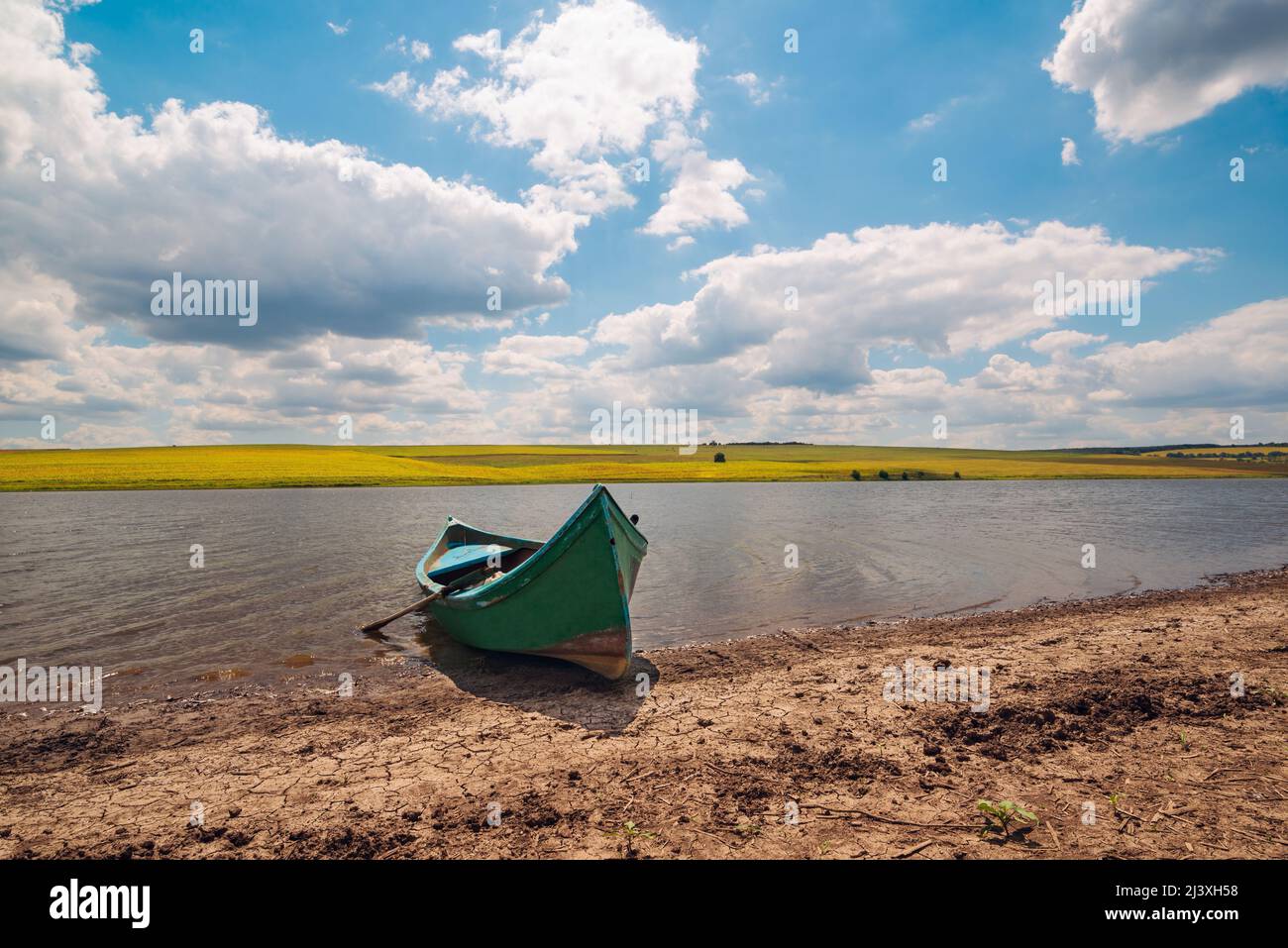 Vieux bateau de pêche sur la rive du lac en face d'un sol brun sec, derrière un beau paysage rural avec des champs jaunes et un ciel bleu avec des nuages. Banque D'Images