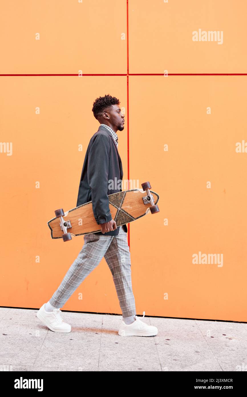 jeune homme avec blazer et skateboard marchant dans la rue sur fond orange. Banque D'Images